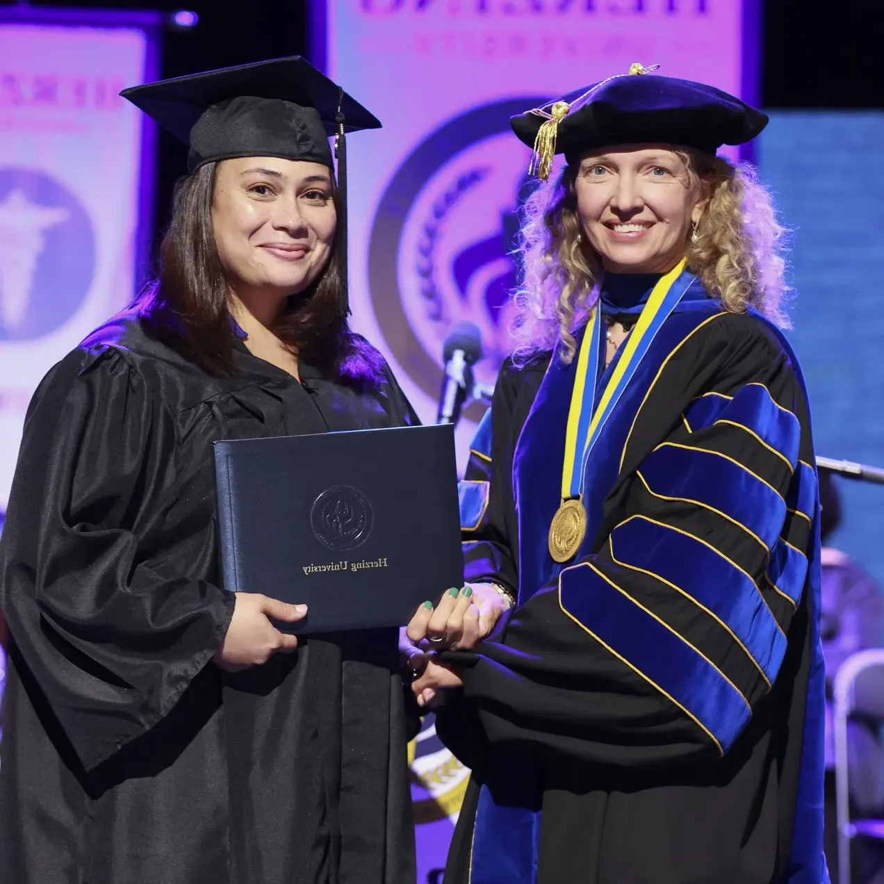 A Herzing University graduate receiving her diploma on stage, accompanied by a faculty member in ceremonial regalia.